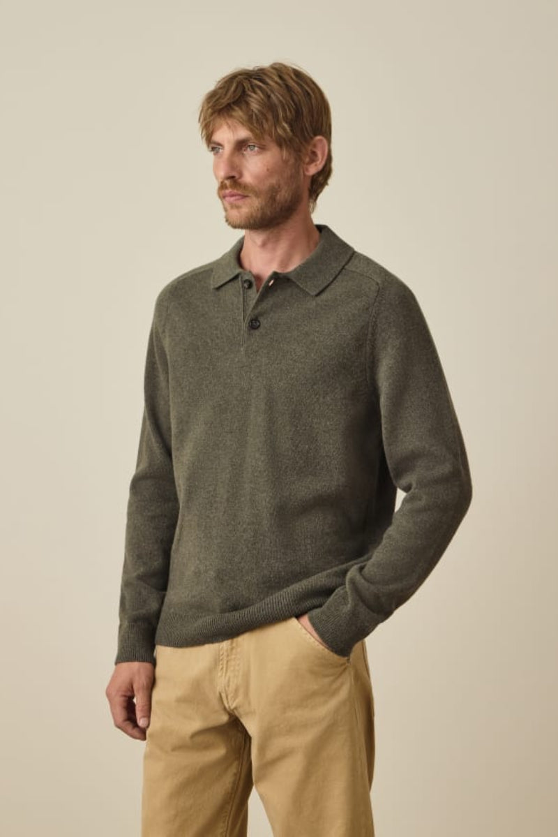 A man wearing a dark olive green long-sleeve knit polo sweater and tan pants, standing against a plain beige studio background.