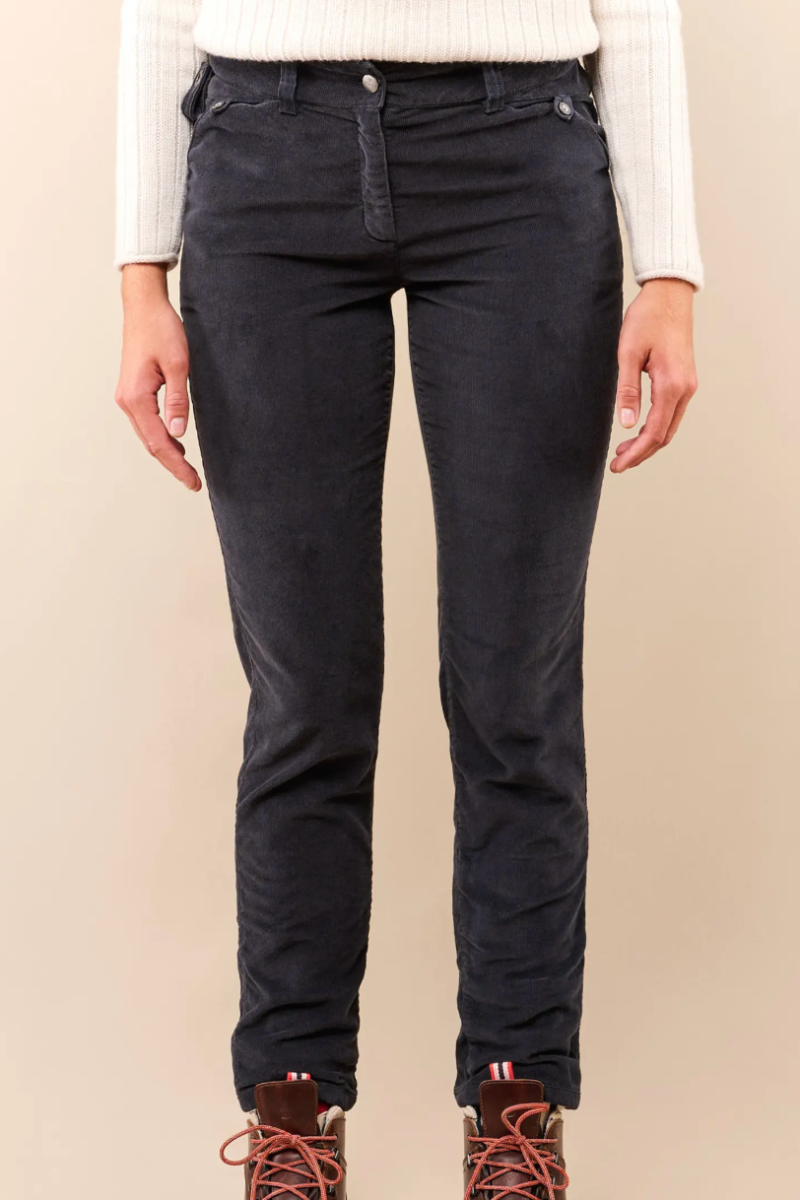 A person wearing slim-fit dark navy jeans, a white ribbed long-sleeve top, and brown lace-up boots, standing against a plain beige studio background.