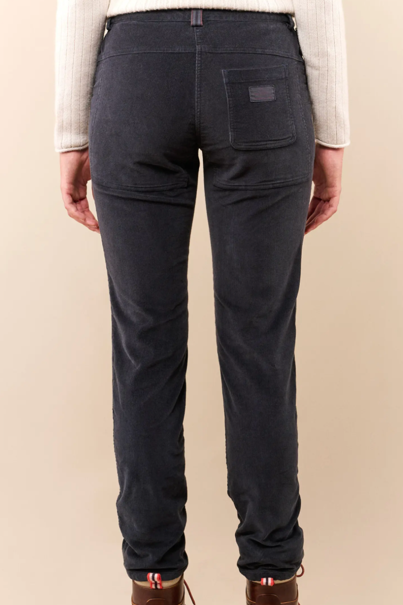 Rear view of a person wearing dark charcoal corduroy pants with a cream knit top and brown lace-up boots, against a plain beige studio background.