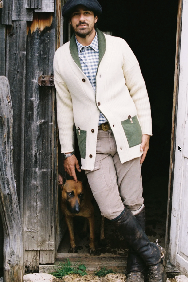 A man wearing a cream ribbed cardigan with green pockets and collar, a blue-and-white check shirt, beige pants, and black boots, standing in a rustic doorway with a brown dog behind him.