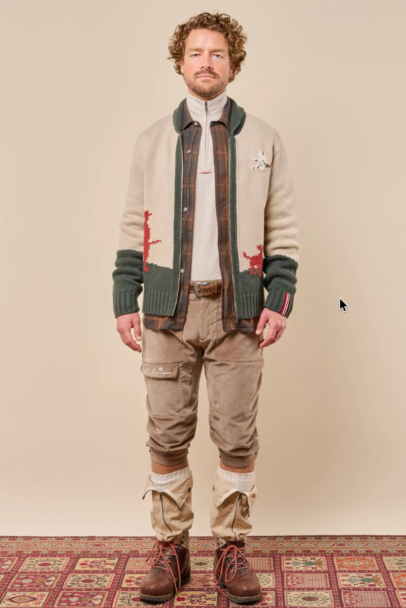 A man stands against a plain beige studio background wearing a cream knit cardigan with dark green ribbed trim and red embroidered details, layered over a brown plaid shirt, white turtleneck, tan cargo pants, and brown lace-up boots.