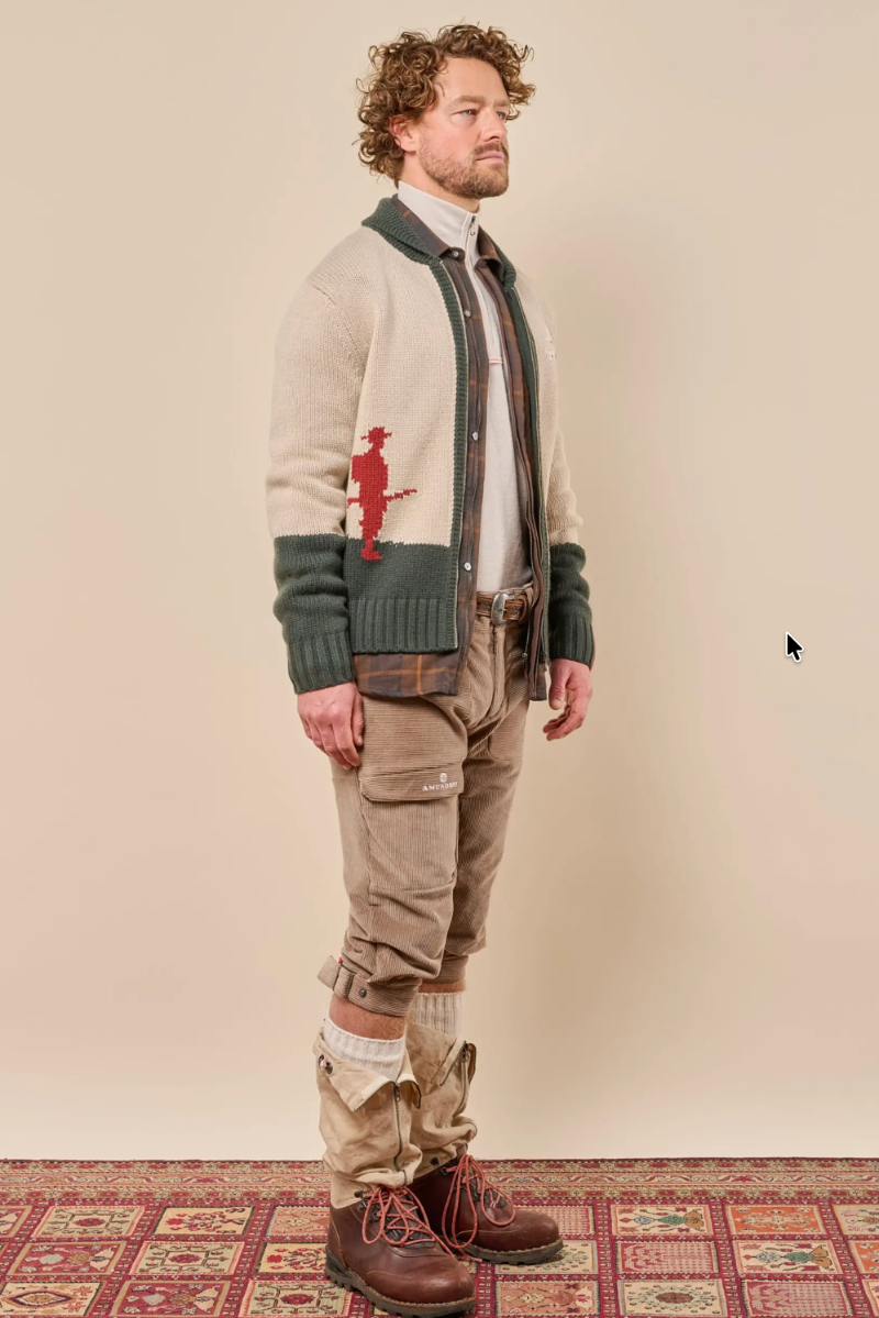 A man stands in profile wearing a cream knit cardigan with dark green trim and a red embroidered figure, layered over a white shirt and brown tie. He wears tan cargo pants, white gaiters, and brown lace-up boots against a plain beige wall and patterned rug.
