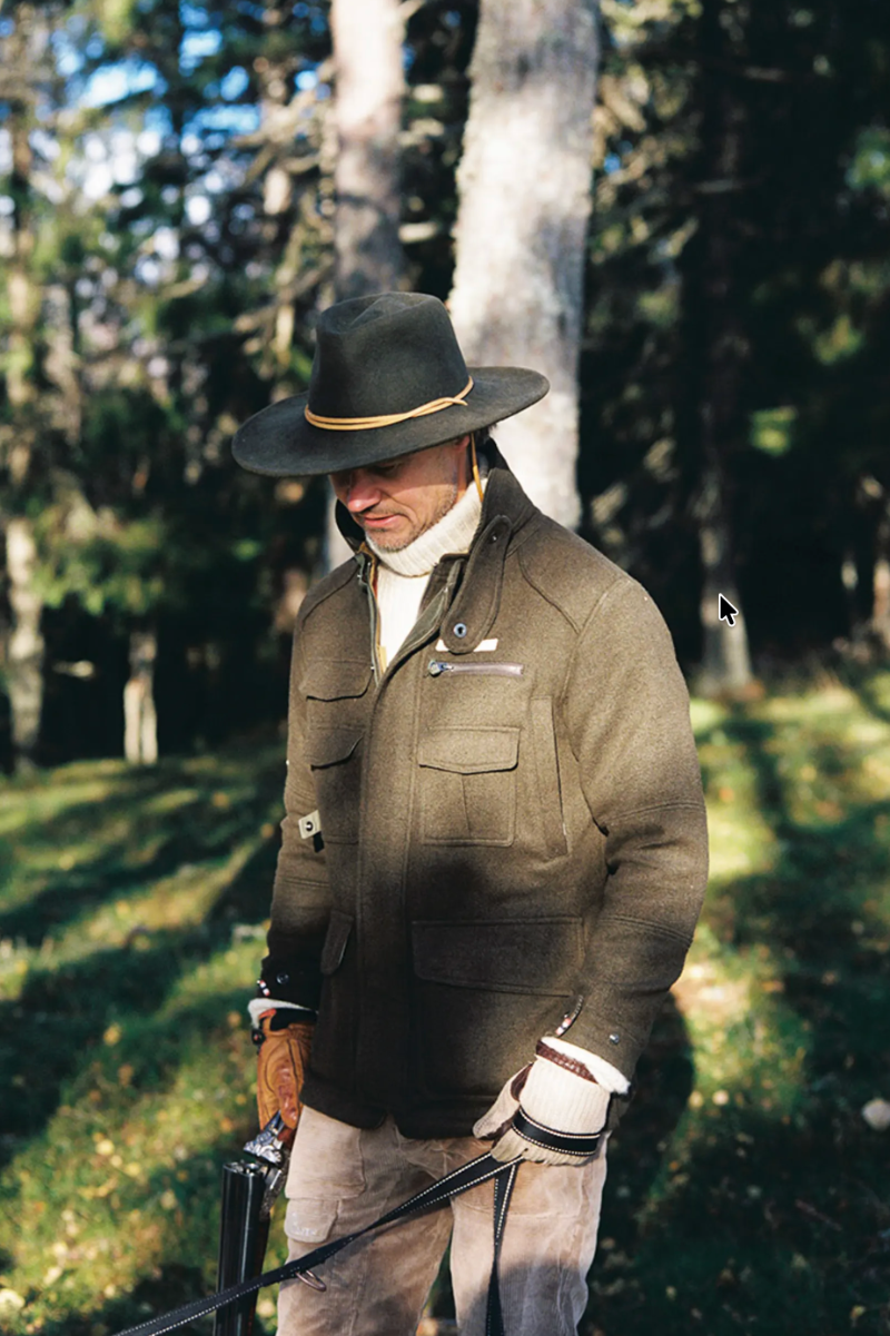 A man wearing a brown wide-brim hat with a yellow band, a brown wool-like jacket, and tan pants stands in a sunlit forest clearing holding a rifle.