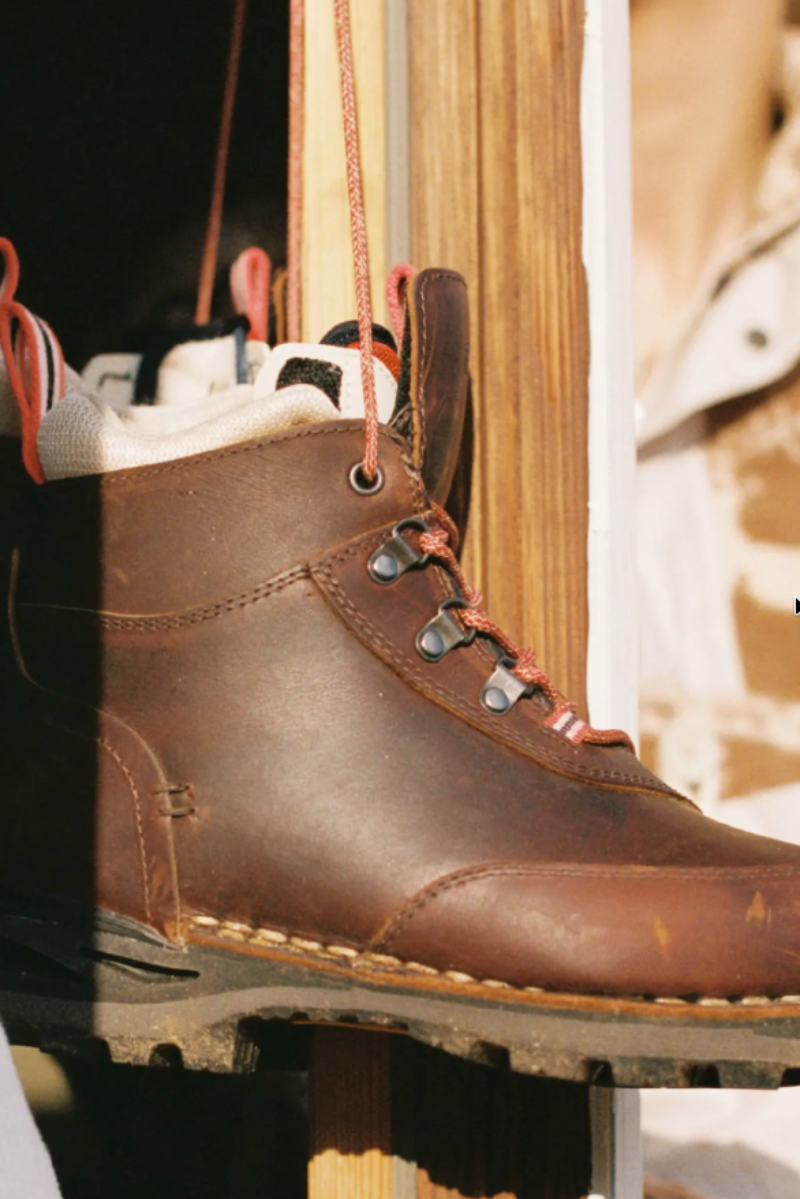 A close-up of a brown leather lace-up boot hanging vertically, with metal eyelets, orange-brown laces, and a rugged sole, set against a warm wooden and neutral background.