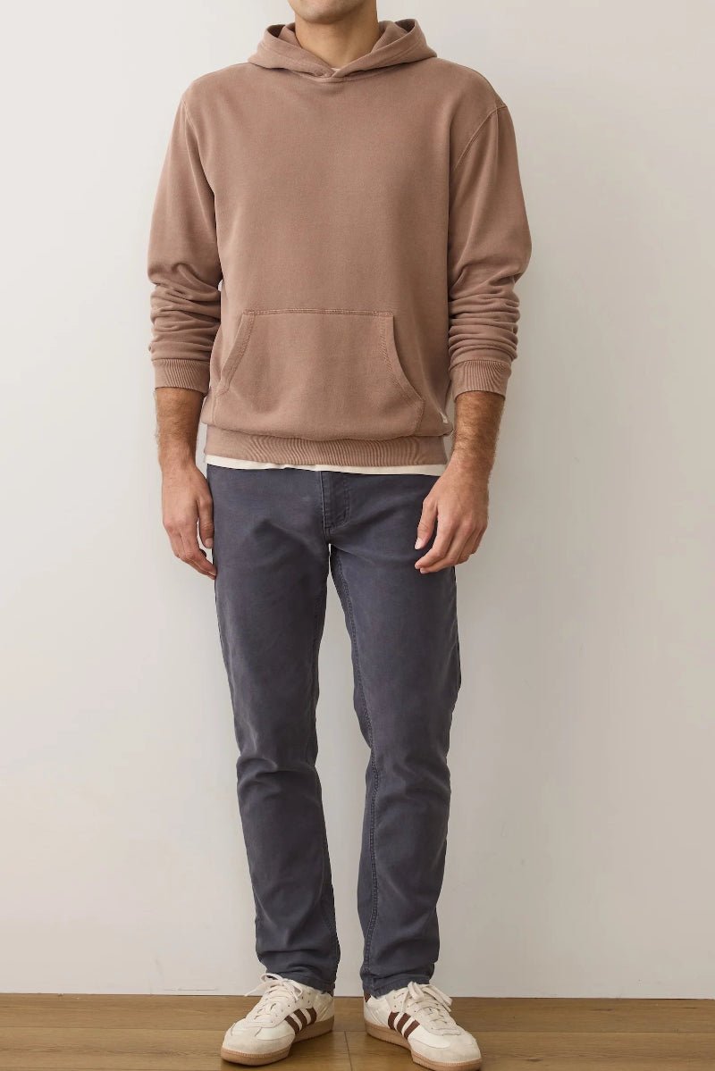 Person wearing a brown hoodie and dark jeans standing against a plain white wall.