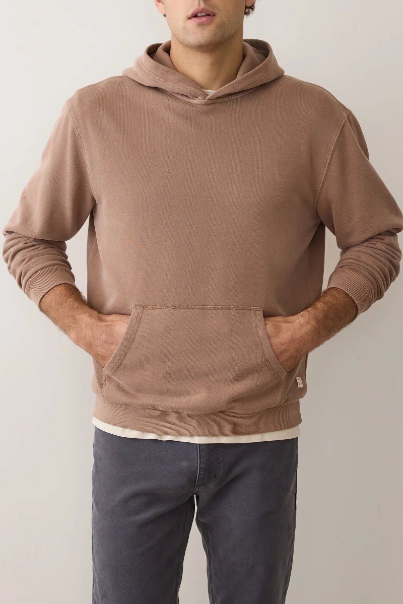 Person wearing a brown hoodie and dark pants against a plain background