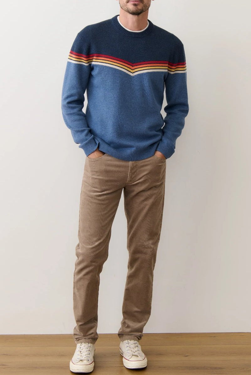 Man wearing a blue sweater with colorful stripes and brown pants against a plain background