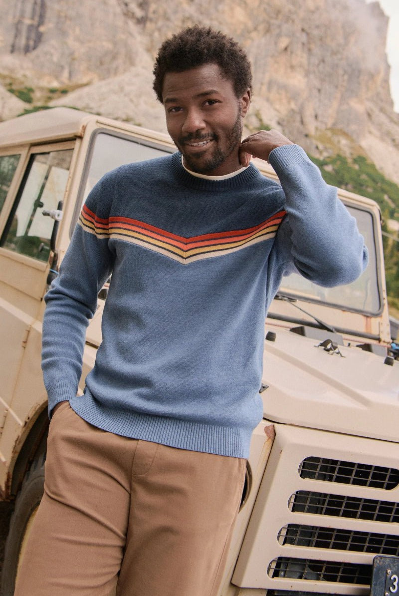 Man in a blue sweater with colorful stripes standing next to a vintage SUV in a mountainous area.