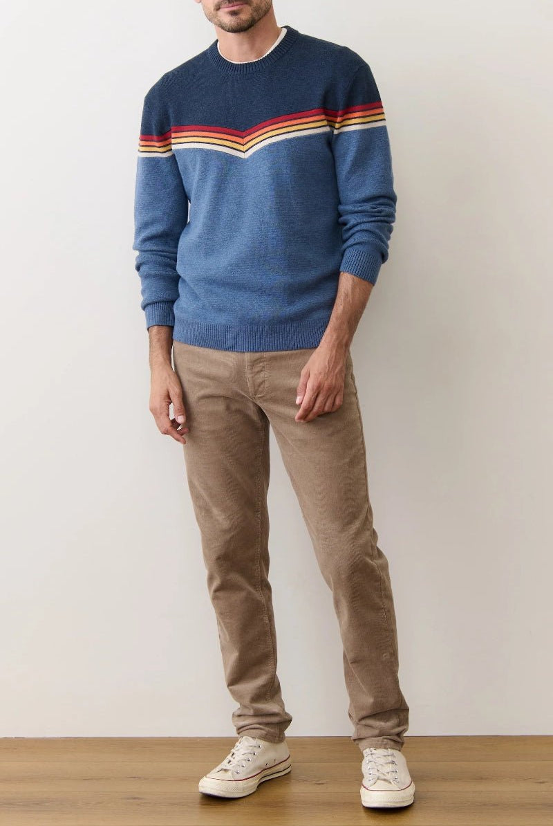 Person wearing a blue sweater with colorful stripes and beige pants against a plain background
