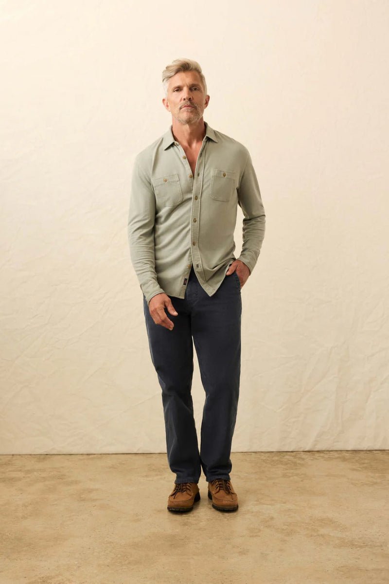 Man wearing a light green shirt, dark blue jeans, and brown boots against a beige wall.