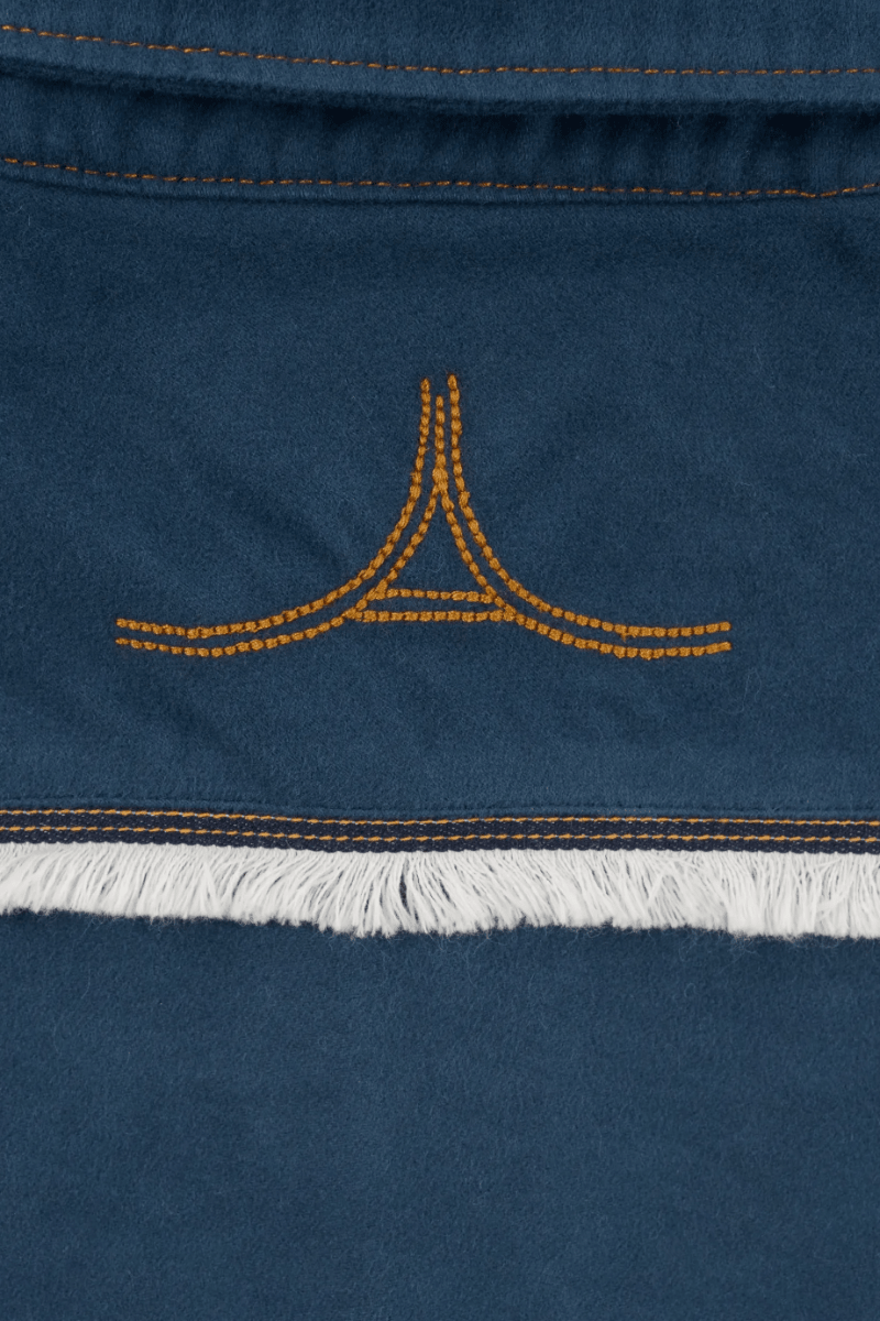 Close-up of a dark blue suede or nubuck textile with orange contrast stitching, a small orange embroidered logo motif, and a white fringe trim.
