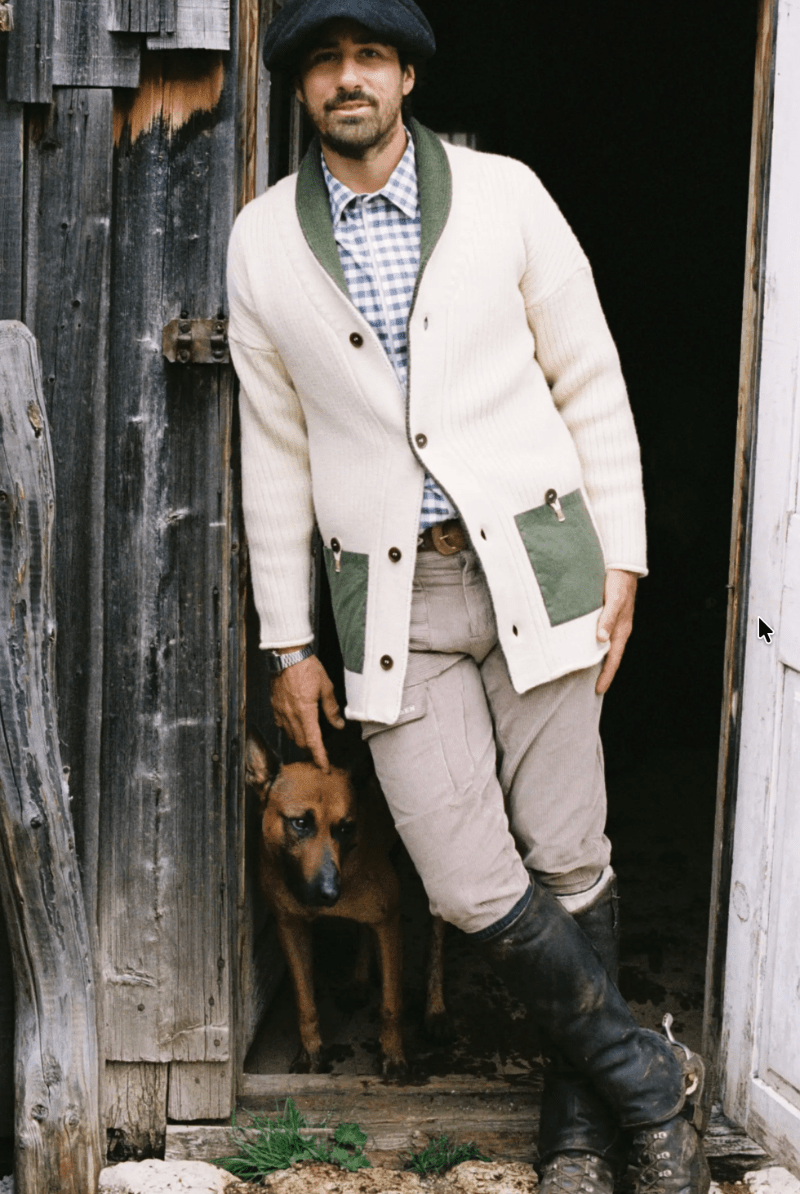 A man wearing a cream ribbed cardigan with green pockets and collar, a blue-and-white check shirt, beige pants, and black boots, standing in a rustic doorway with a brown dog behind him.