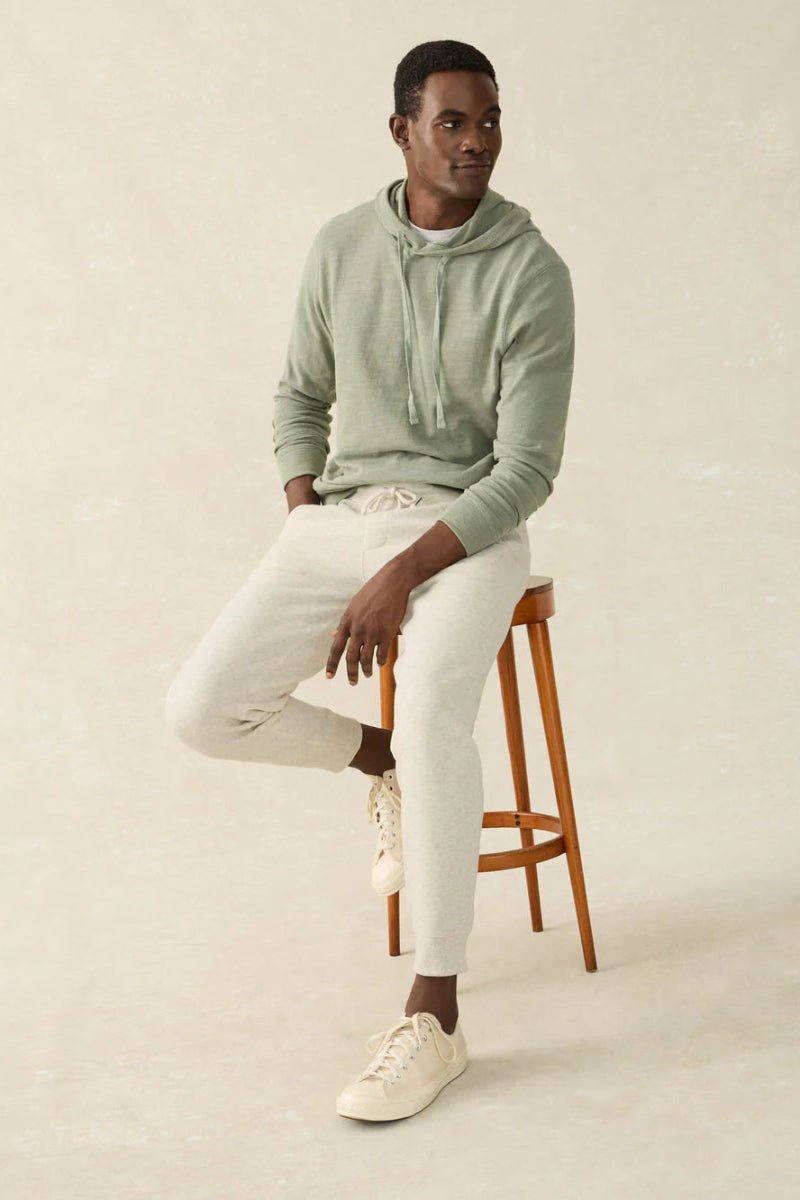 Man sitting on a wooden stool wearing a light green hoodie and white pants against a beige background