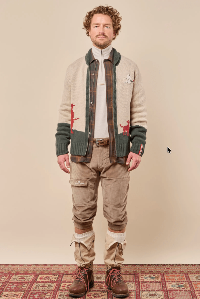 A man stands against a plain beige studio background wearing a cream knit cardigan with dark green ribbed trim and red embroidered details, layered over a brown plaid shirt, white turtleneck, tan cargo pants, and brown lace-up boots.