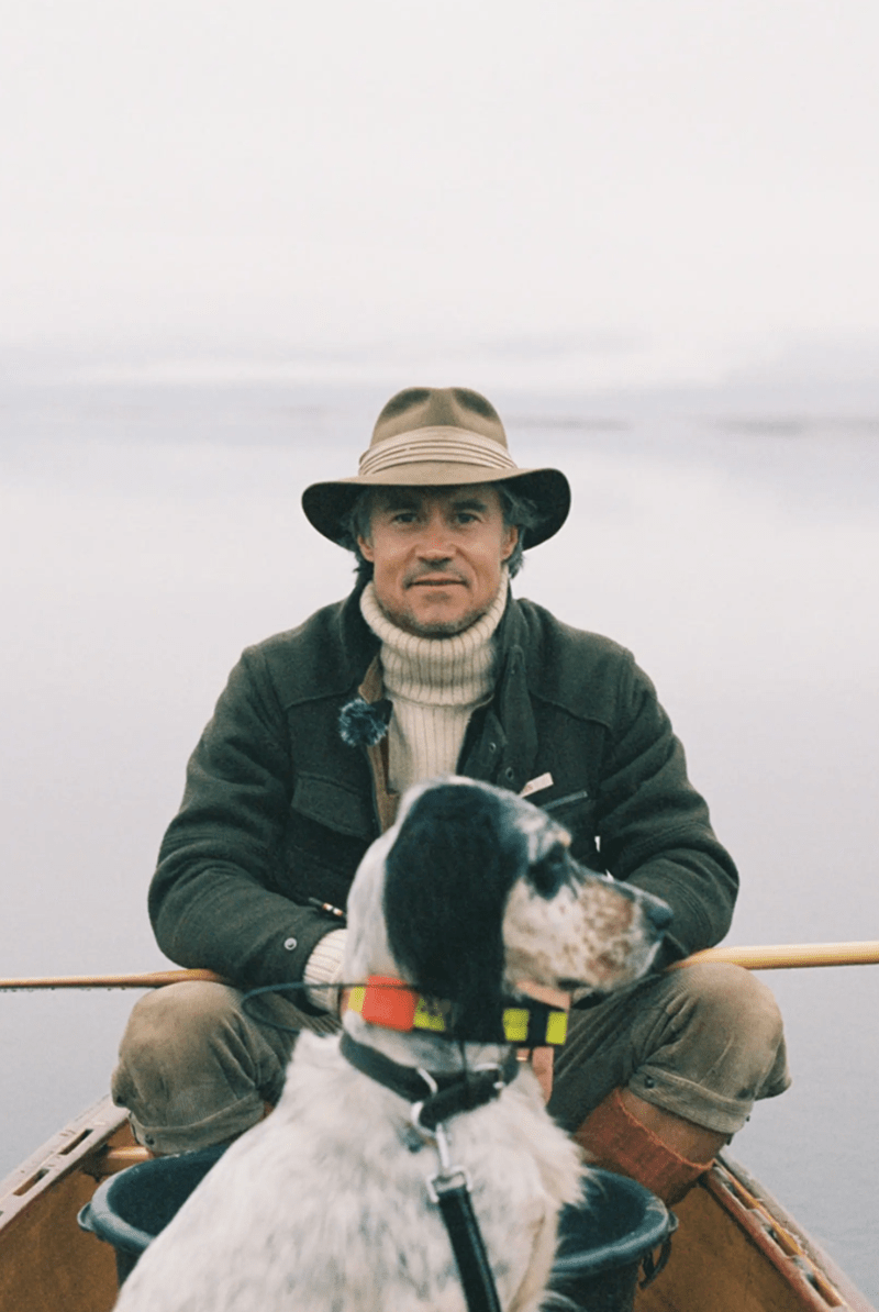 A man wearing a wide-brimmed hat, dark jacket, and cream turtleneck sits in a wooden boat with a black-and-white dog on a lake under an overcast sky.
