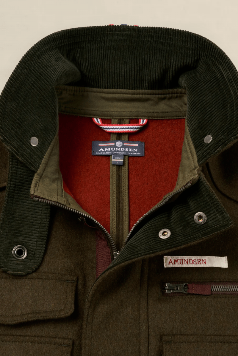 Close-up of an Amundsen jacket showing the interior collar, olive green corduroy lapels, red fleece lining, and a sewn-on brand label on the chest pocket.