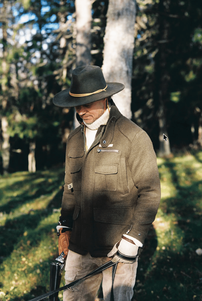 A man wearing a brown wide-brim hat with a yellow band, a brown wool-like jacket, and tan pants stands in a sunlit forest clearing holding a rifle.