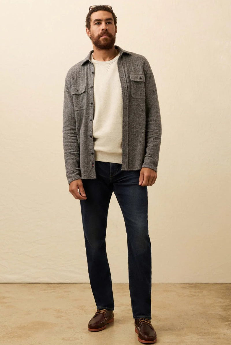 Man wearing a gray checkered shirt over a white t-shirt, dark jeans, and brown shoes against a plain background.