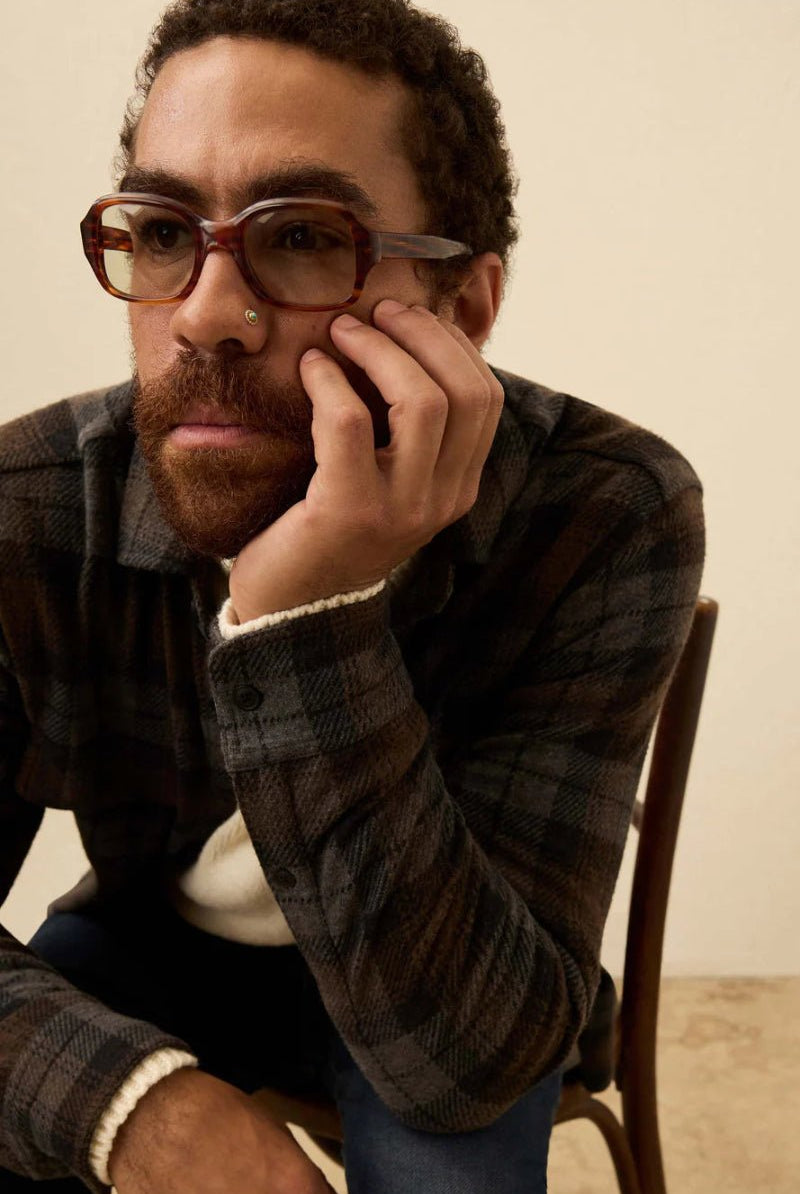 Man wearing glasses and a plaid shirt sitting on a chair against a beige wall.