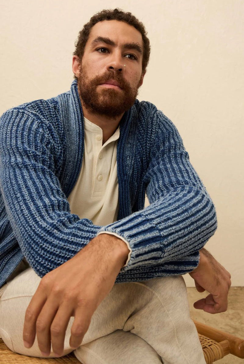 Man wearing a blue knitted cardigan over a white shirt, sitting on a wooden chair.