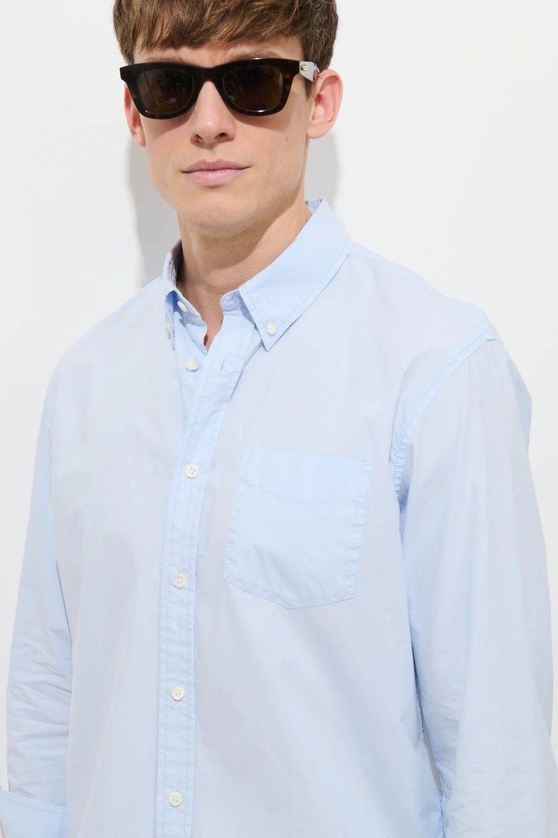 Man wearing a light blue button-up shirt and sunglasses against a white background