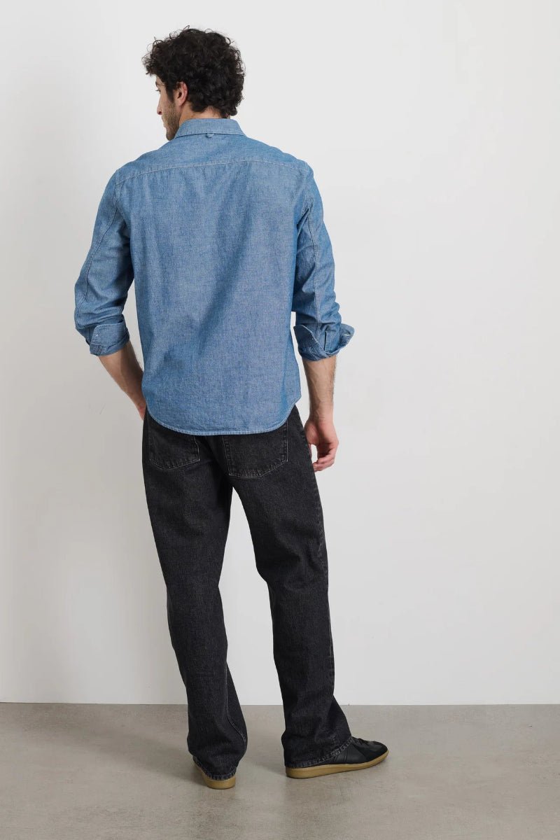 Man wearing a blue denim shirt and black jeans standing against a white wall.