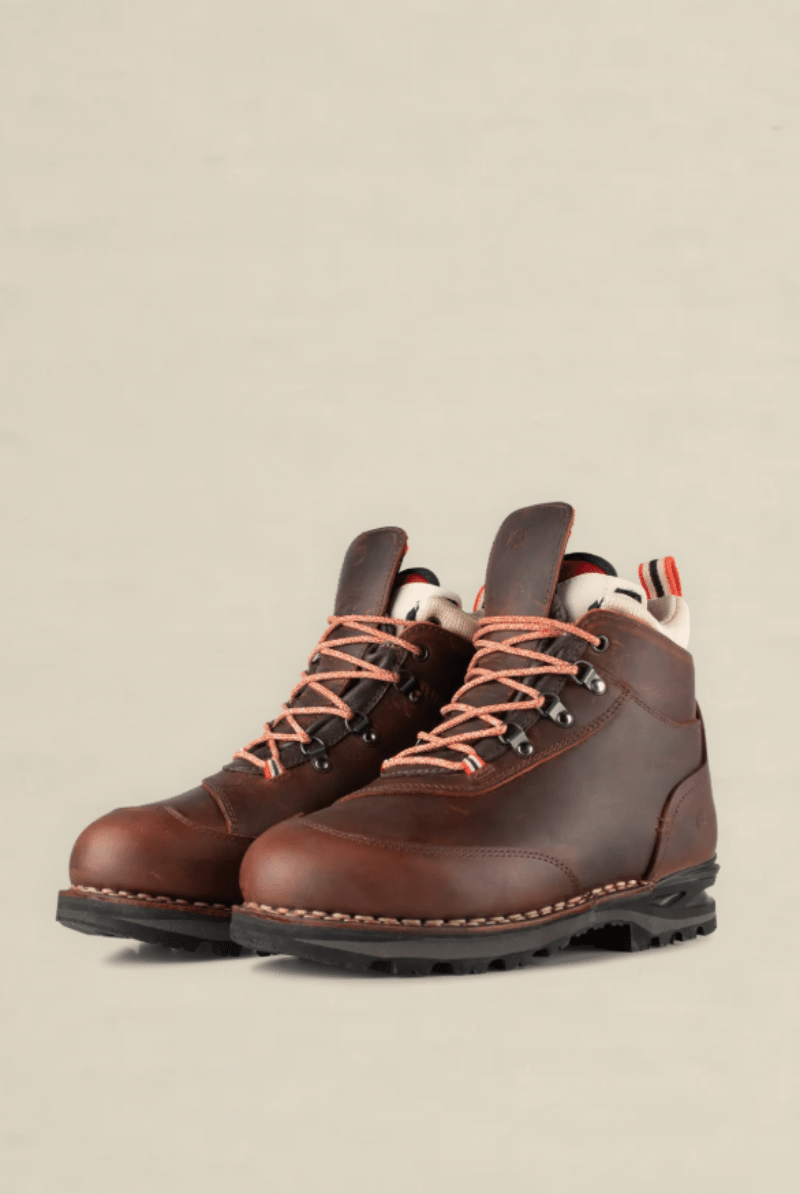 A pair of brown leather hiking boots with orange laces and a black sole, photographed on a clean, solid beige background.