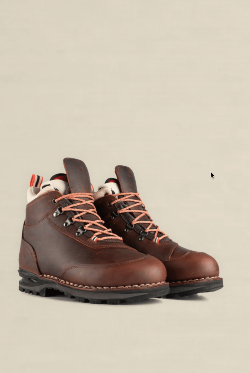 A pair of brown leather hiking boots with orange laces and a cream lining, shown against a solid beige background.