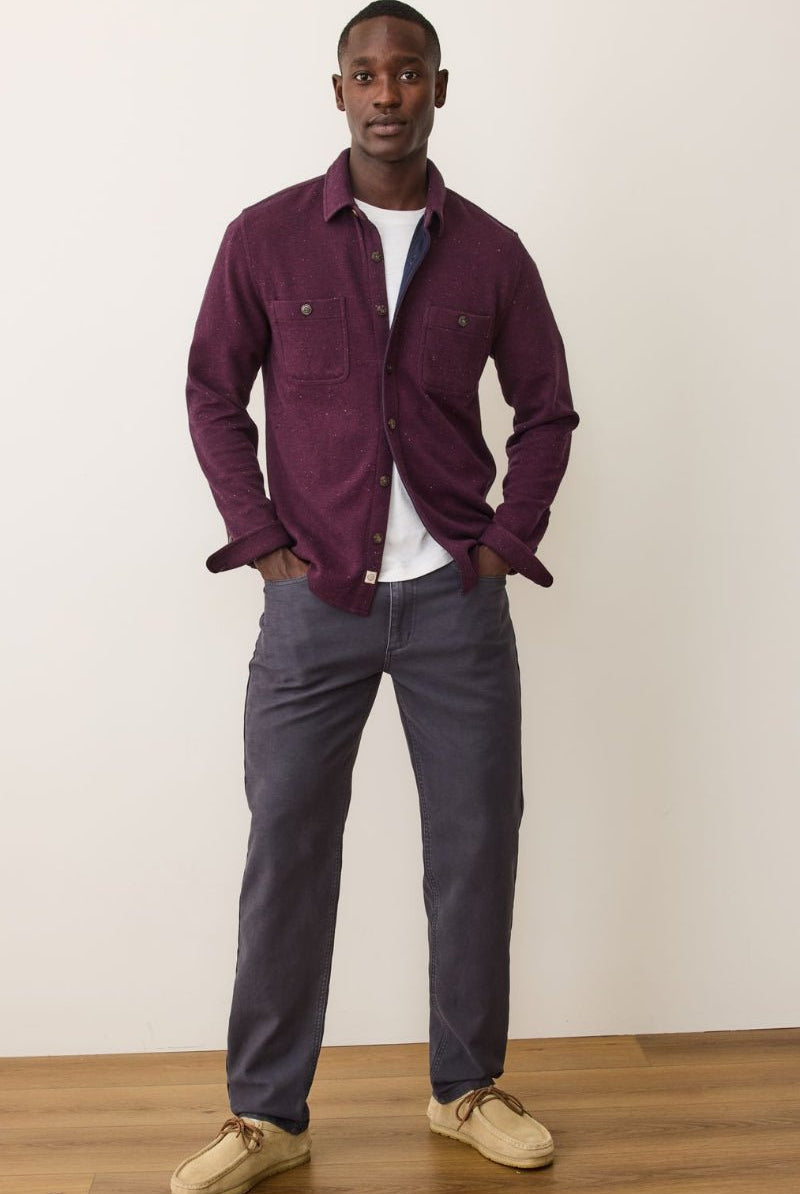 Man wearing a purple shirt and gray pants standing against a white wall.