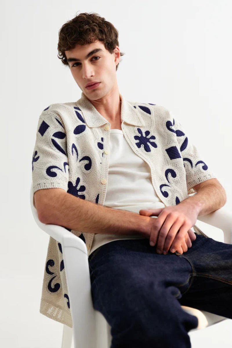 Man wearing a patterned cardigan sitting on a white chair against a plain background