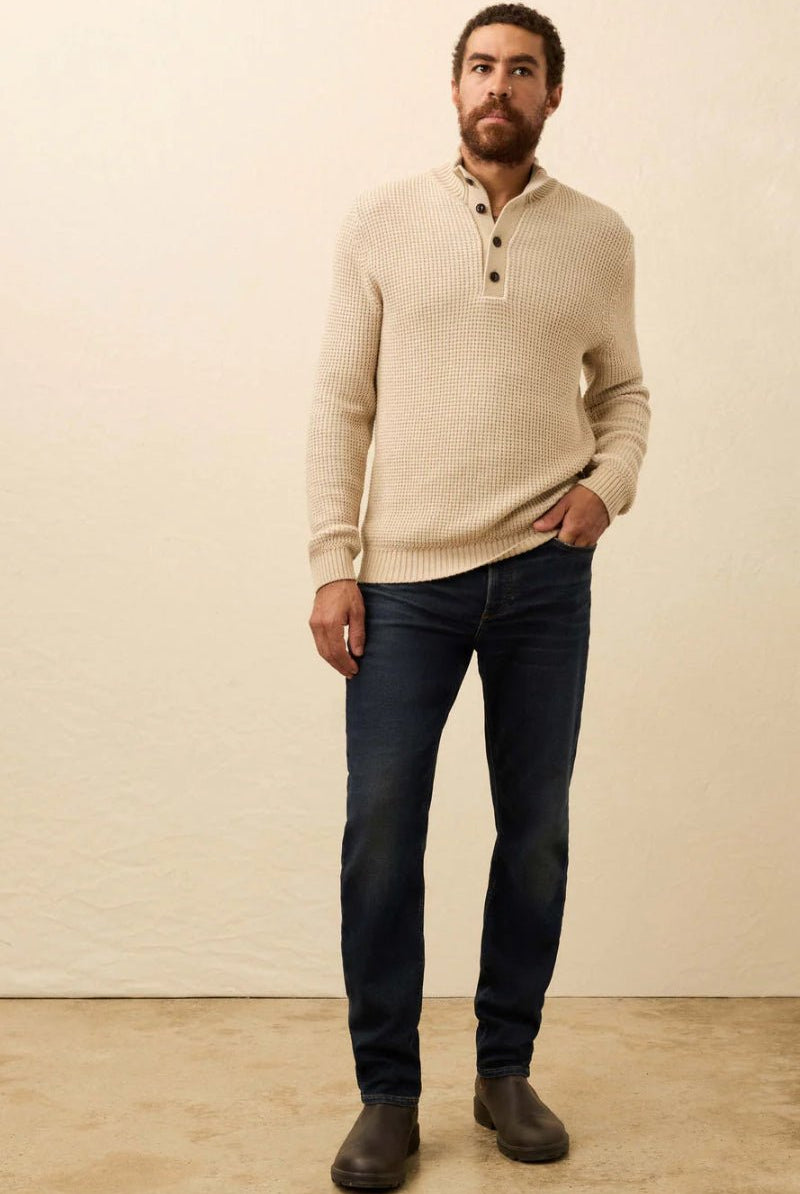 Man wearing a beige sweater and dark jeans against a plain background