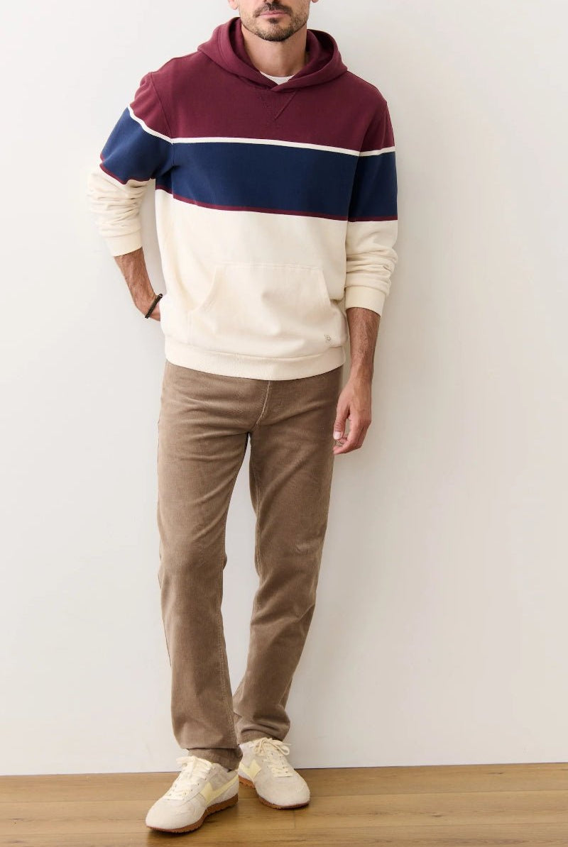 Person wearing a color-blocked hoodie and brown pants against a white wall.