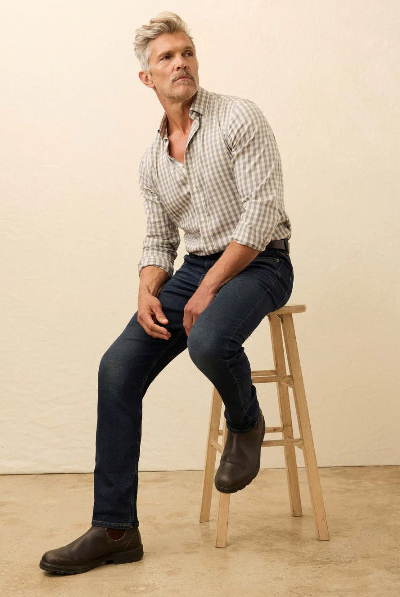 Man sitting on a wooden stool wearing a checkered shirt and dark jeans against a beige background