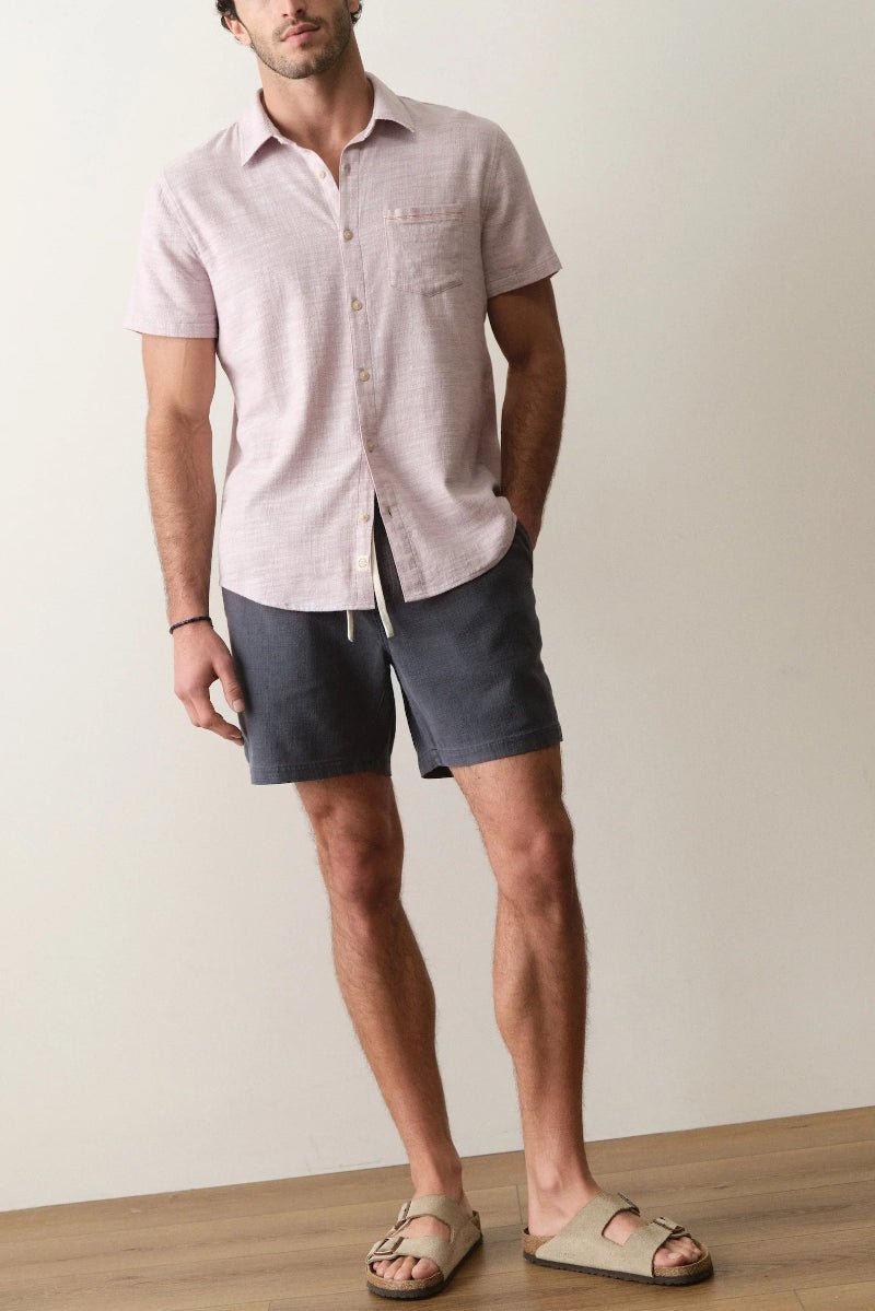Man wearing a light pink short-sleeve shirt and dark shorts against a plain background