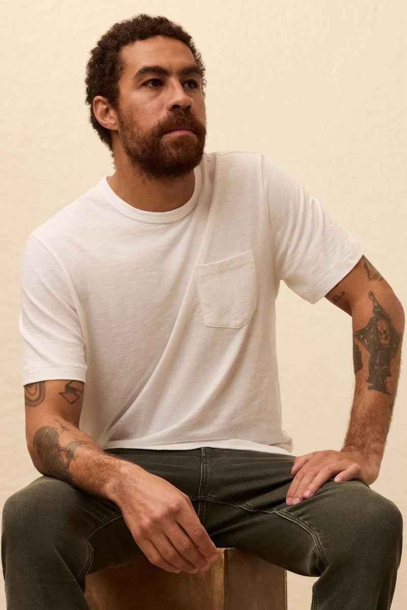 Man wearing a white t-shirt and dark jeans sitting on a wooden stool against a beige background