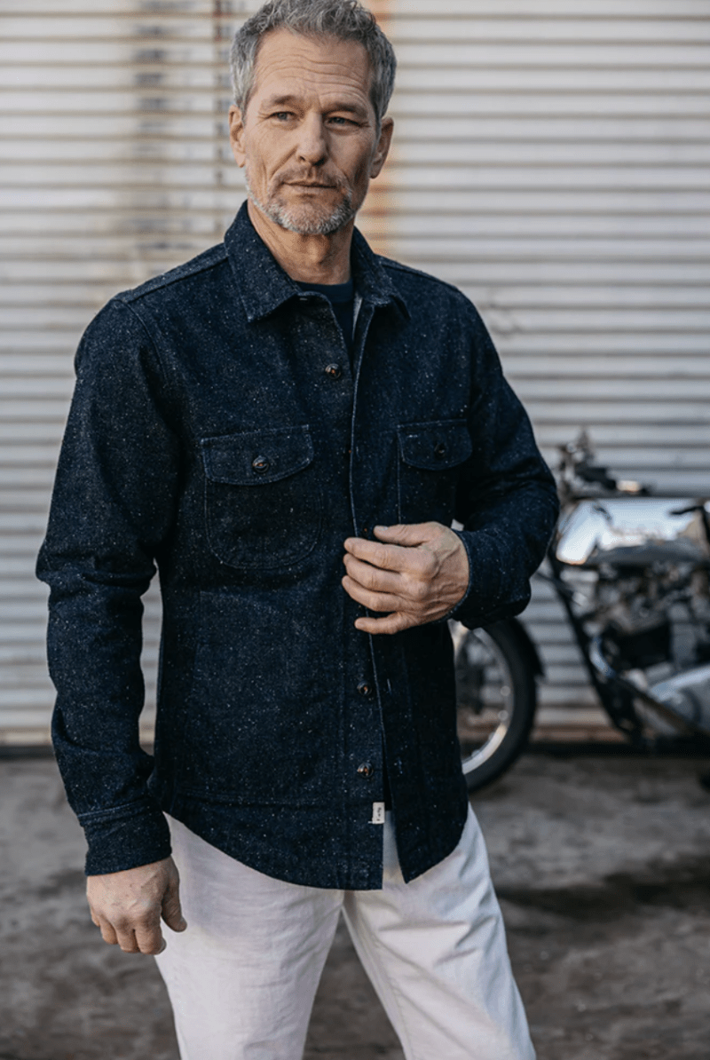 Man wearing a dark blue jacket and white pants standing in front of a motorcycle.
