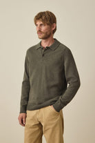A man wearing a dark olive green long-sleeve knit polo sweater and tan pants, standing against a plain beige studio background.