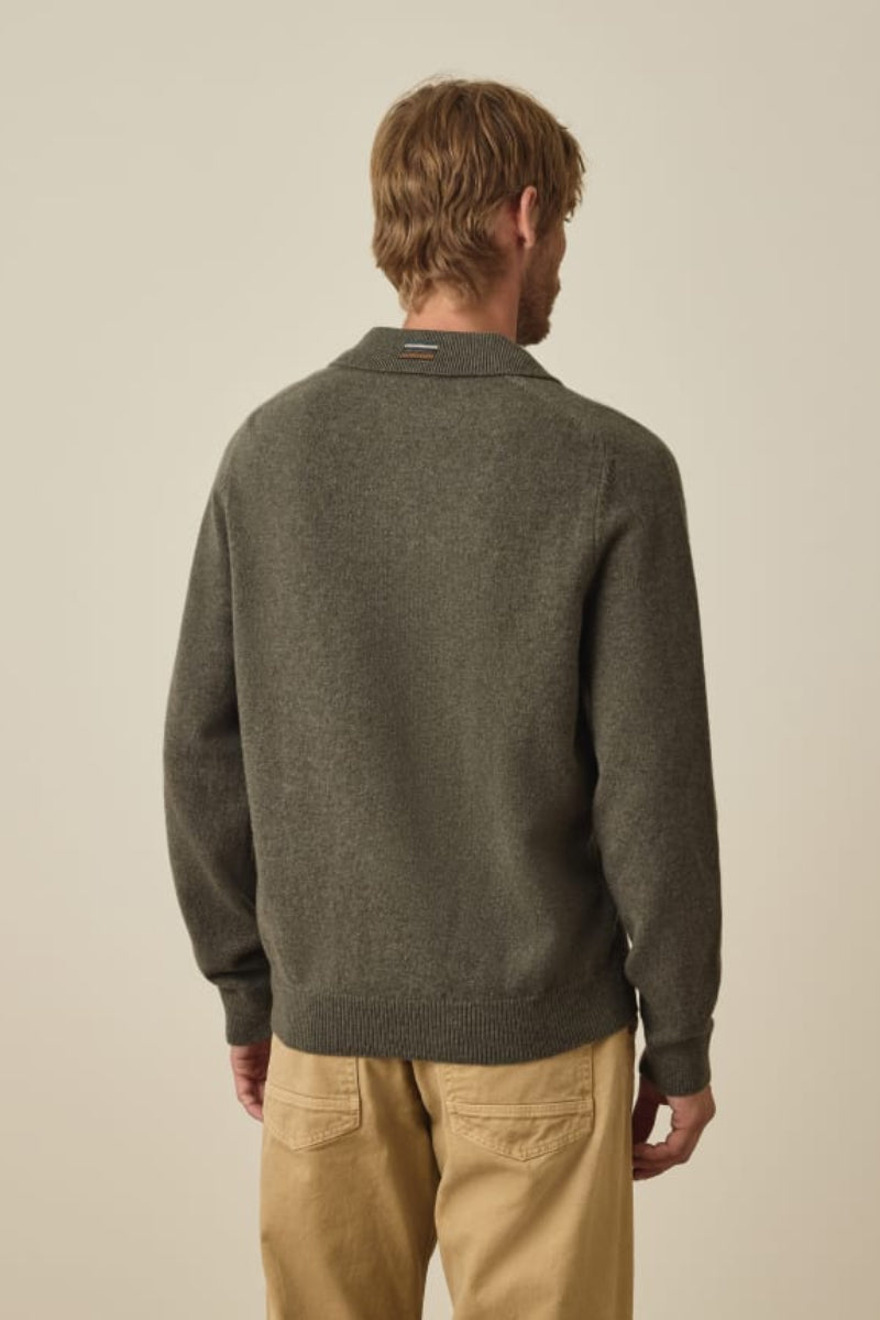 A man viewed from behind wearing a dark olive green knit sweater and tan pants, standing against a plain beige studio background.