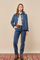 A woman stands against a plain beige studio backdrop wearing a blue denim shirt and matching jeans, layered over a white turtleneck, with a brown leather belt and brown lace-up boots. A patterned rug is visible at the bottom.