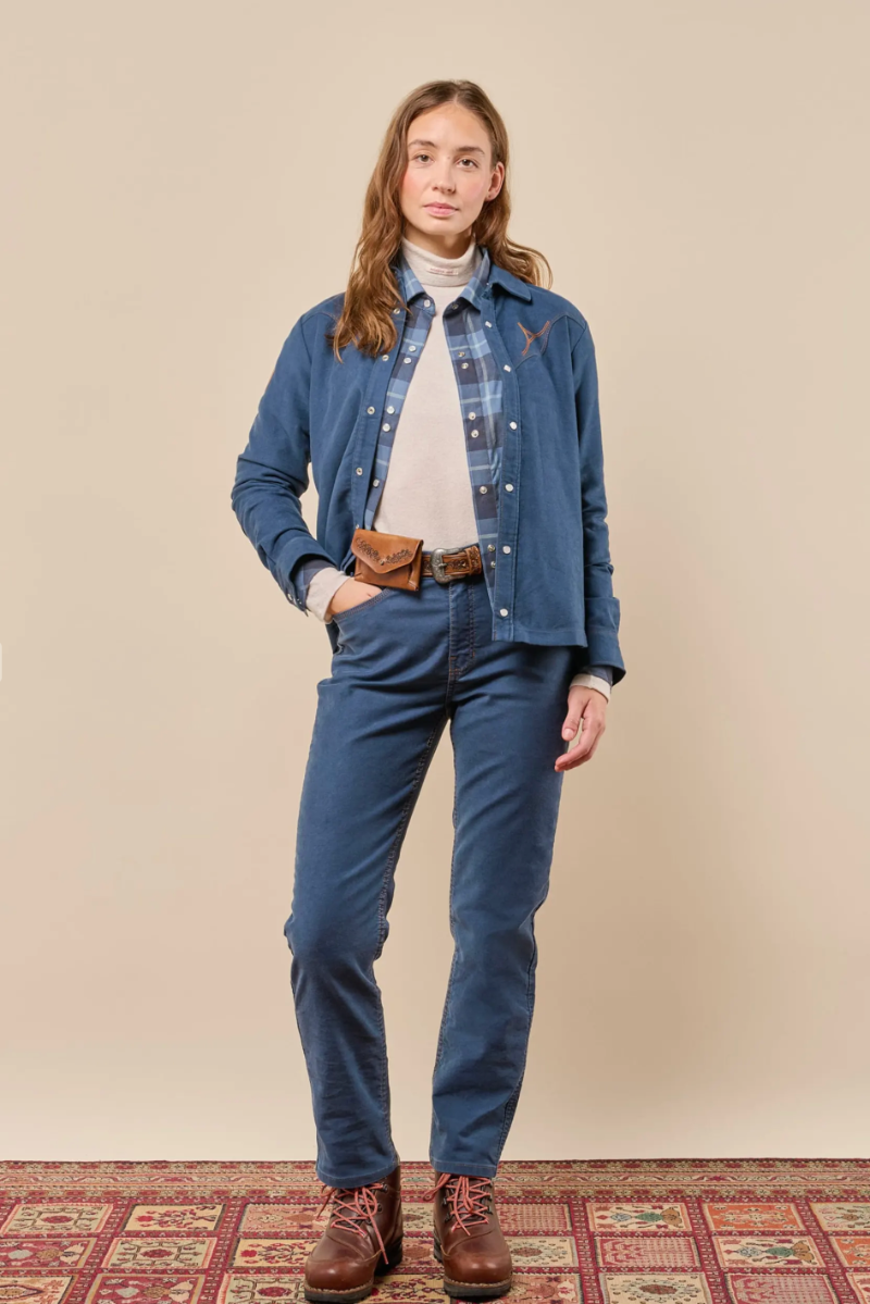 A woman stands against a plain beige studio backdrop wearing a blue denim shirt and matching jeans, layered over a white turtleneck, with a brown leather belt and brown lace-up boots. A patterned rug is visible at the bottom.