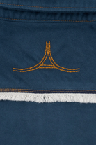 Close-up of a dark blue suede or nubuck textile with orange contrast stitching, a small orange embroidered logo motif, and a white fringe trim.