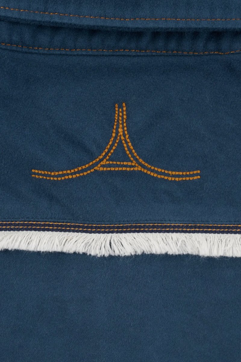 Close-up of a dark blue suede or nubuck textile with orange contrast stitching, a small orange embroidered logo motif, and a white fringe trim.