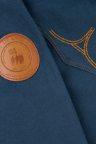 Close-up of a dark teal wool-blend jacket featuring a circular brown leather patch on the chest with embossed text and a gold embroidered geometric pattern.