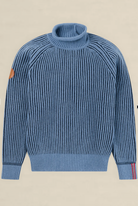 A blue ribbed knit turtleneck sweater with a brown leather patch on the left sleeve and thin red and white stripes on the right sleeve cuff, shown flat against a light beige background.