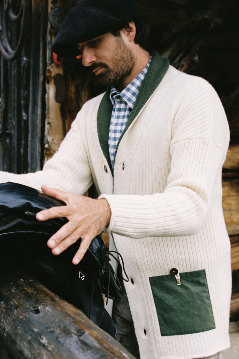 A man wearing a cream ribbed cardigan with dark green trim and a green patch pocket, layered over a blue gingham shirt and black beret, leaning on a rustic wooden log and touching a black bag.