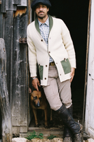 A man wearing a cream ribbed cardigan with green pockets and collar, a blue-and-white check shirt, beige pants, and black boots, standing in a rustic doorway with a brown dog behind him.