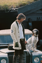 A man wearing a cream knit cardigan over a white shirt and dark trousers stands beside a dog on the hood of a vintage blue vehicle in front of a rustic wooden structure.