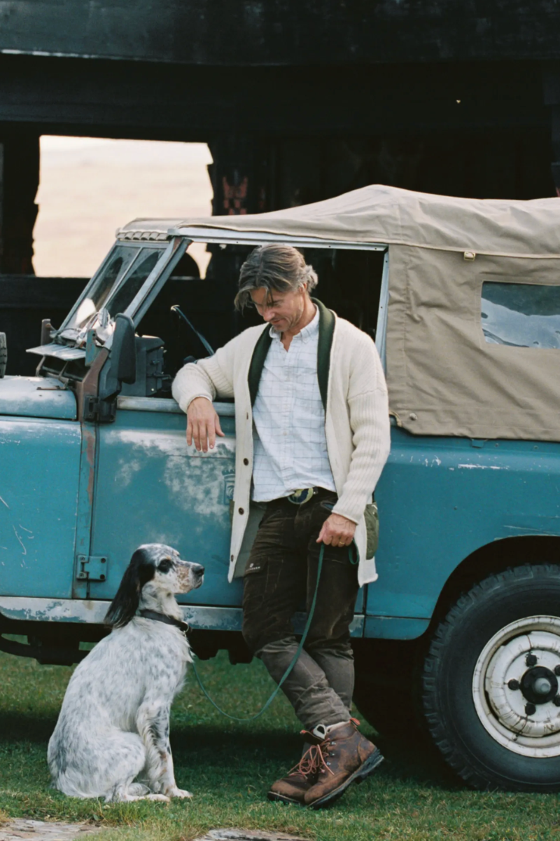 A man in a cream cardigan and brown boots leans against a vintage blue off-road vehicle next to a white and black spotted dog on a grassy outdoor setting.