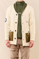 A man wearing a cream ribbed cardigan with olive green panels, a green half-zip turtleneck, and beige cargo pants, photographed against a plain beige studio background.