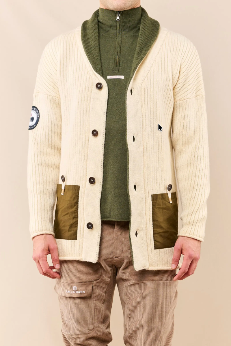 A man wearing a cream ribbed cardigan with olive green panels, a green half-zip turtleneck, and beige cargo pants, photographed against a plain beige studio background.